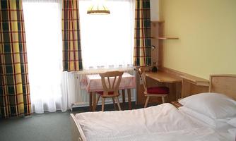A simple bedroom with a double bed, a desk, and a chair. The windows are decorated with checked curtains and let in plenty of light.