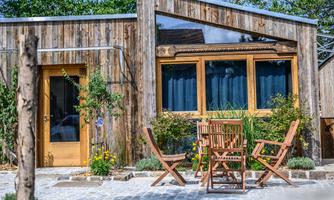 A modern wooden house with large windows and a green surrounding. In front of the house is a cozy table with chairs.