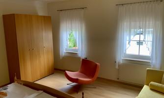 A cozy bedroom with a wooden box and large windows. A red chair and a sofa provide a modern accent in the room.