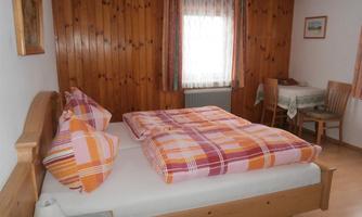 A cozy bedroom with wooden decor. The bed has striped blankets and there is a small dining area in the background.