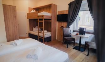 A cozy room with two beds, a bunk bed, and a table. There are large windows and modern wooden furniture.