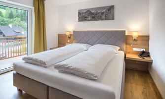 A modern bedroom with a double bed and light wooden floor. The large window front offers a view of the nature outside.
