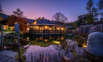 A quiet pond with a lit building surrounded by trees. The sky is in soft colors of the sunset.