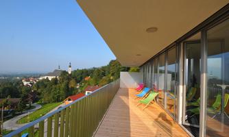 A modern terrace with colorful loungers and a view of a picturesque landscape. In the background, a church and green hills can be seen.