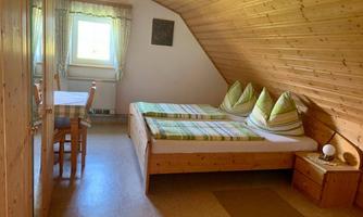 A cozy room with a wooden ceiling and two beds. A table with chairs and a window provide bright lighting.