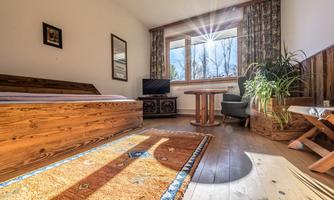 A cozy room with a wooden bed and a window through which sunlight shines. There is a table, a chair, and a plant that create an inviting atmosphere.