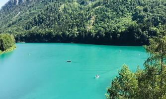 A clear, turquoise lake surrounded by lush green forest and mountains. Some boats can be seen on the water.