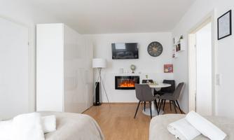 A modern living room with a television and an electric fireplace. There is a dining table with chairs and a bright, welcoming atmosphere.