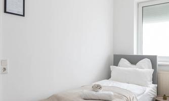 A bright bedroom with a single bed and cozy bedding. The room has white walls and a large window with natural light.