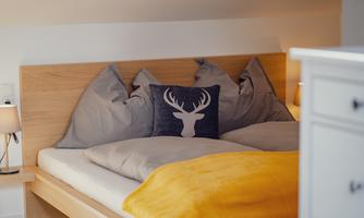 A cozy bed with gray pillows and a decorative pillow featuring a deer motif. The blanket is yellow and the room has a warm, inviting atmosphere.