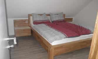 A cozy bedroom with a double bed and two pillows. The floor is tiled with wood, and there is a small nightstand.