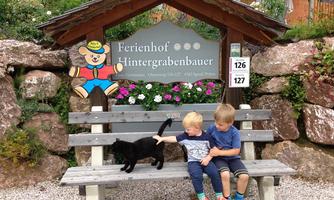 Two children are sitting on a bench and petting a black cat. In the background, a sign with the name "Ferienhof Hintergrabenbauer" is visible.