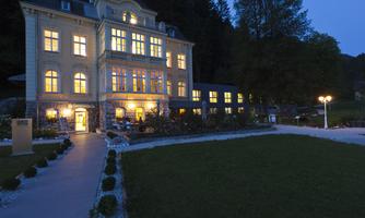 A beautiful, illuminated building in the evening light, surrounded by a well-maintained garden. Trees in the background give the scene a tranquil atmosphere.