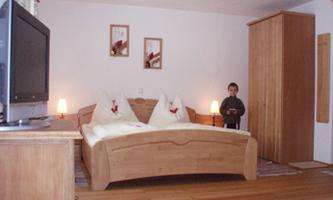 A cozy bedroom with a large wooden bed and two heart-shaped pillows. In the room, a television and a wardrobe can be seen.