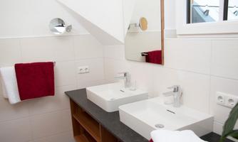 A modern bathroom with two sinks and large mirrors. Red towels and a wooden shelf give the room an inviting atmosphere.