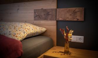 A cozy bedroom with a wooden bed and a colorful pillow. A small vase with dried flowers is on the bedside table.