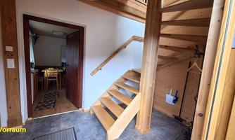 A bright hallway with a wooden staircase and a cozy dining area in the background. The walls are plain and the floor is tiled.