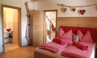 A cozy bedroom with a double bed and red pillows. The walls are made of wood, and the room is cheerful with heart-shaped decorations.