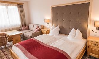 A cozy bedroom with a large bed and a padded headboard. The room is furnished with a sofa and wooden furniture and has plenty of natural light.