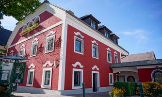 A red building with white stucco and several windows. Surrounded by well-maintained plants and a clear blue sky.