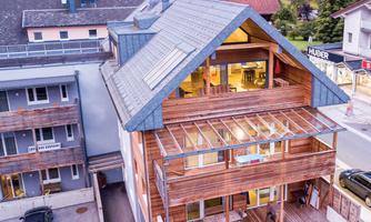 A modern wooden house with large windows and a sloped roof. It is located on a quiet street, surrounded by other buildings.