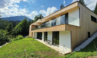 A modern house with wooden cladding and large windows in a green environment. In the background, mountains and a blue sky can be seen.