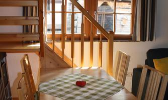 A bright and cozy dining area with a wooden table and chairs. In the background, there is a staircase and windows with curtains.