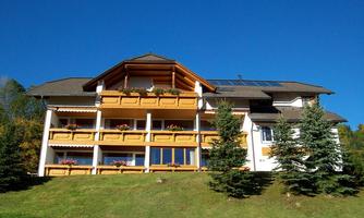 A modern building with a balcony and flower boxes. The sky is clear and blue, surrounded by green trees.