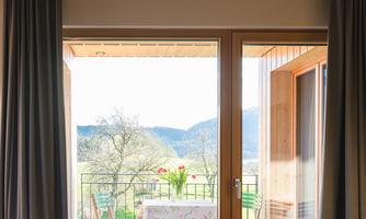 A cozy living space with a large window and a view of a terrace. On the table are flowers and in the background, green hills are visible.