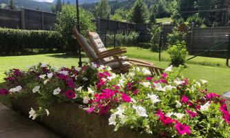A beautiful garden with colorful flowers and a deck chair. In the background, green trees and mountains can be seen.