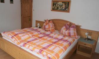 A cozy bedroom with a wooden bed and colorful pillows. The walls are plain, and there is a door as well as small pictures in the background.