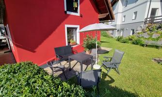 A cozy outdoor area with a table and chairs in a well-kept garden. In the background, there is a red house.