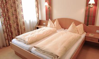 A cozy bedroom with two single beds and bright, beige bedding. The walls are adorned with floral curtains and decorative lamps.