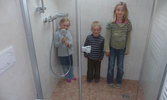 Three children are standing in a modern shower. The wall tiles are bright and the shower is glass-enclosed.