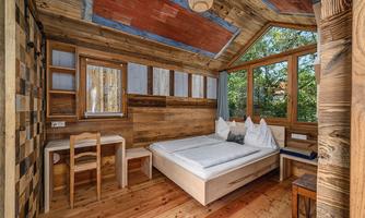 A cozy bedroom with wood decor and large windows. There is a bed, a table, and wooden chairs, surrounded by plants.