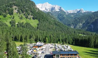 A picturesque mountain landscape with green forests and snow-capped peaks. In the foreground, there is a parking lot and a building in nature.
