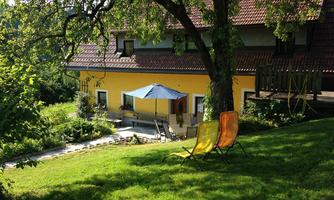 A cozy house with a yellow facade and a garden. Two lounge chairs are situated on the green grass under a tree.