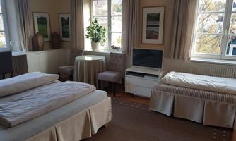 A cozy room with two beds and a small table. Large windows let in plenty of light, and there are plants and pictures on the walls.