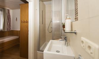A modern bathroom with a shower and a sink. In the background, a sleeping area with a wooden wardrobe is visible.