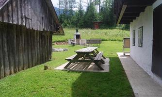 A cozy meadow with a wooden table and benches. In the background, wooden huts stand and trees frame the scene.