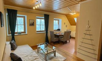 A cozy room with wooden floor and wooden beam ceiling. There is a sofa, a table, as well as a dining area with chairs and windows with curtains.