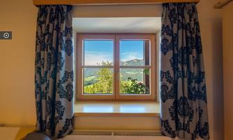 A window with two wings, surrounded by decorative curtains. Outside, there is a landscape with mountains and fresh greenery.