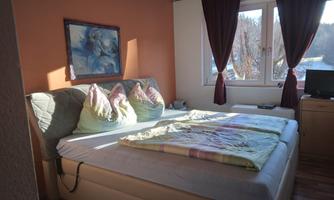 A cozy bedroom with a large bed and colorful cushions. The windows let in plenty of daylight and offer a view outside.