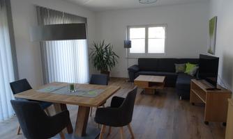 A modern living room with a wooden dining area and comfortable chairs. The room has large windows that let in plenty of light.