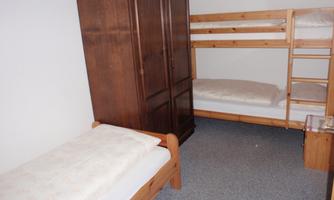 A simple room with two beds, a bunk bed, and a wardrobe. The floor is carpeted, and the walls are plain.