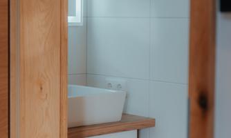 A modern bathroom with a white sink and wooden shelves. The walls are light-tiled and the light streams through a window.