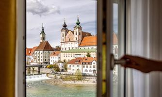 A view from a window of a picturesque town with historic buildings and a church. The river flows gently below, surrounded by colorful houses.