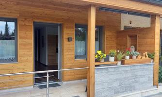 A modern wooden house with an inviting entrance area. In front of the house, there are blooming plants in pots.