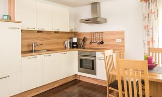 A modern kitchen with white cabinets and wooden shelves. Four wooden chairs are at the table and there is a cooking area with appliances.