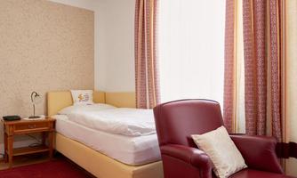 A simple room with a single bed and a red armchair. The windows are bright and the walls are designed with a subtle pattern.